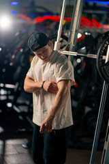 Fototapeta na wymiar in the gym young handsome pumped up guy in a T-shirt and cap stands looking at his biceps