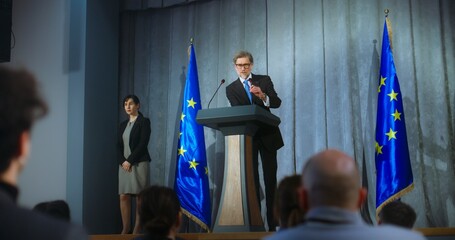 Mature politician comes to tribune, delivers campaign speech, answers journalists questions, gives interview to media. Representative of the European Union performs at press conference. Elections day. - obrazy, fototapety, plakaty