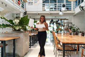 cheerful African woman entrepreneur or owner holds paper in spacious cafe with modern decor. joyful expression and casual relaxed, creative atmosphere, possibly reviewing positive business outcome..