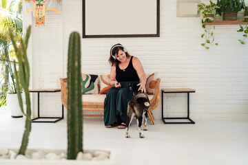 cheerful middle-aged Caucasian woman with headphones enjoys time with dog in stylish, plant-filled indoor setting. sitting on rattan bench, touching small, black dog, exuding relaxation and joy
