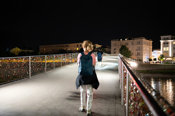 Salzburg, Austria, August 15, 2022. Iconic night image of the pedestrian bridge with padlocks placed by tourists in love. A middle-aged woman is walking along it.