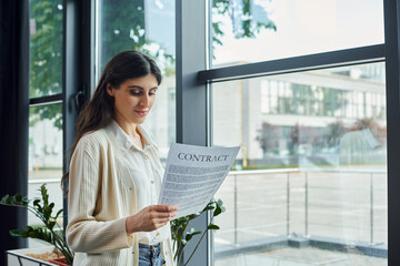 A modern businesswoman reads a contract while standing by a window in her office, surrounded by a...