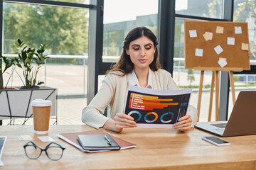 A businesswoman sits at a table, engrossed in charts in a modern office setting.