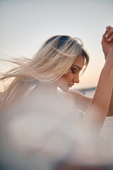 A young blonde woman facing away, hair flowing in the breeze during a picturesque sunset The warmth...