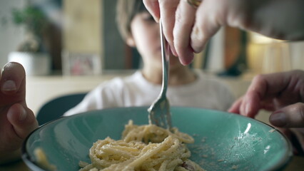 Close-up of fork spinning spaghetti on blue plate with little boy in blurred background, mother's...