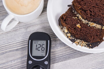 Glucose meter with result sugar level, sweet chocolate cake and cup of coffee with milk. Measuring and checking sugar level during diabetes