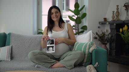 Happy pregnant woman showing ultrasound picture of baby while seated on couch sofa at home during...