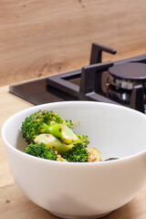 The white bowl with stir-fried broccoli on the wooden table near the gas stove