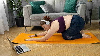 Sporty plus size woman with headphones in sportswear working out at home. Sports, activity and weight loss concept. Young woman in leggings exercising on the mat, watching online video via laptop
