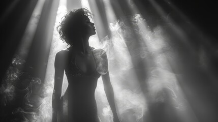   A black-and-white image of a woman, hands on hips, silhouetted against a beam of light