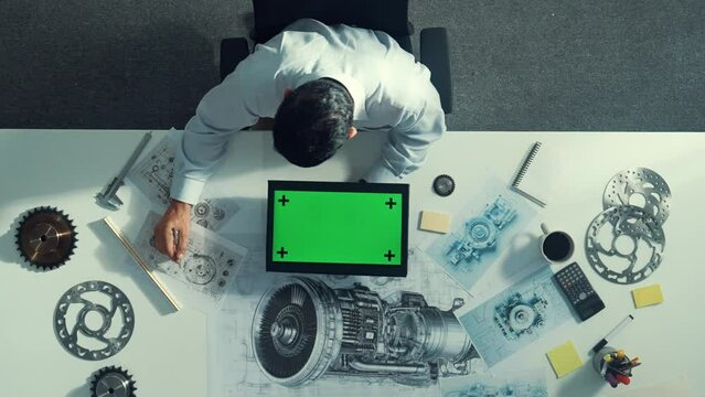 Top view of skilled engineer writing in paper while looking at tablet display green screen with turbine engine sketch design and metal gear and architectural equipment scatter around. Alimentation.