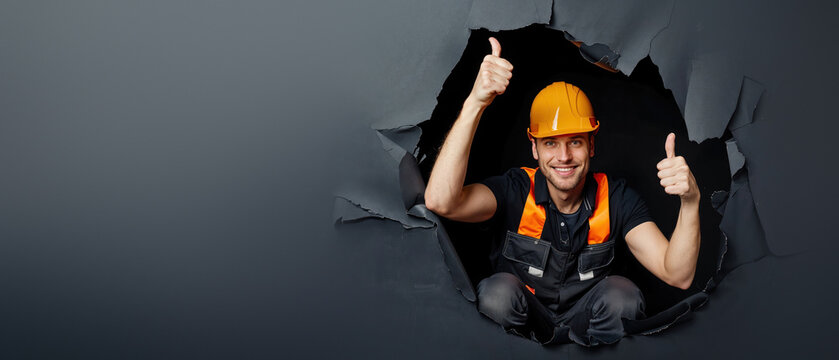 Cheerful builder poses with thumbs up, popping through a dark paper tear, showing confidence