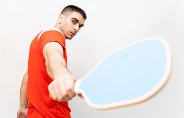 A shaved man in his 30s poses on a white background with a pickleball racket in sports clothing. The young man is backhanding the racket towards the camera. Pickle ball concept.