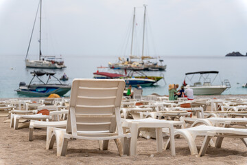 Close-up of white plastic sunbeds on the beach against the background of the blurred sea, mountains, yachts and boats.