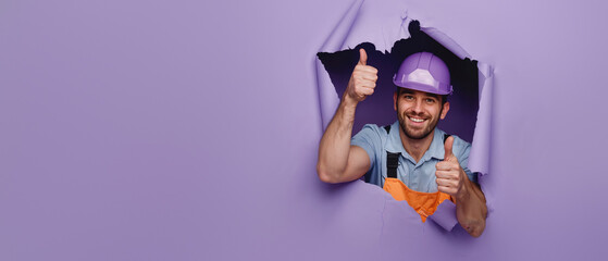 A cheerful construction worker with hard hat giving thumbs up as he breaks through a purple paper wall