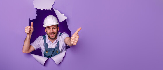 Smiling man with yellow helmet gives thumb up through torn purple paper
