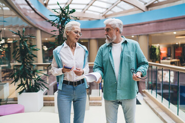 Senior Caucasian couple in casual wear discussing over papers and a smartphone in a shopping mall. Aged man and woman in their 60s appear confused or concerned, indicating a problem-solving 