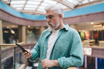 Mature Caucasian man with a white beard in a teal shirt holding glasses and a smartphone in a shopping mall. Male in his 60s looks focused, possibly reading or texting, standing alone indoors