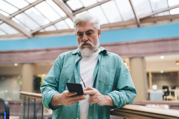 Concentrated elderly Caucasian man with beard using smartphone app in bright office. The senior, in his 60s, seems absorbed by cellular, reflecting tech-savvy generation against indoor modern backdrop