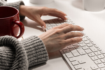 A woman in a sweater is sitting at a computer and typing an email on a white keyboard, working from...