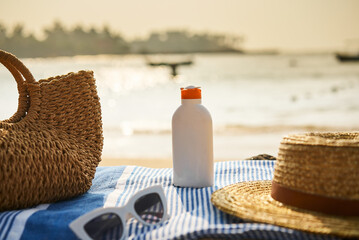 Beach scene with sun cream, hat, sunglasses, and straw bag on towel. Bottle of SPF lotion ready for...
