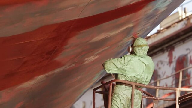 Worker paints hull in shipyard, maintenance of large vessel underway. Protective gear worn for safety as spray gun applies red coating. Maritime industry, ship renovation process showcased in action.