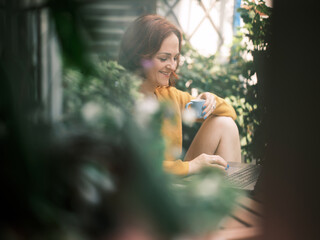 Joyful woman in yellow sweater holding a cup and using laptop in a garden-like balcony setting