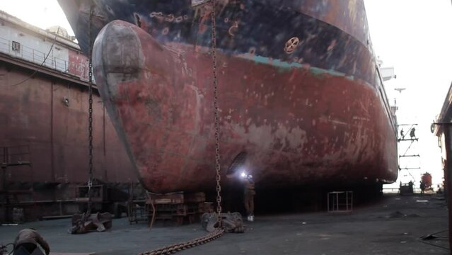Workers repair ship hull in dry dock. Welding, grinding performed on massive vessels bow. Industrial maritime maintenance, shipbuilding process captured. Rust, repair tools, visible in shipyard.