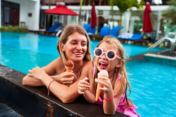 Cheerful young girl with sunglasses enjoys ice cream beside smiling mom in a pool. Relaxing family...