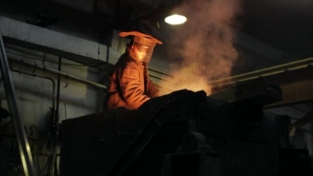 Industrial worker in protective gear oversees molten metal pouring furnace at steel mill. Foundry professional in heat-resistant suit controls smelting process, crucial for manufacturing sector.