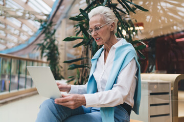 Elegant senior woman with silver hair, using a laptop, focused and engaged, seated in a bright mall, technology literacy in the elderly. Mature businesswoman using laptop computer in offce