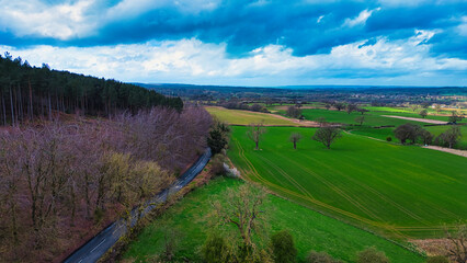 Fototapeta na wymiar Aerial view of a vibrant rural landscape with a road running alongside a forest, contrasting green fields and a dramatic cloudy sky.