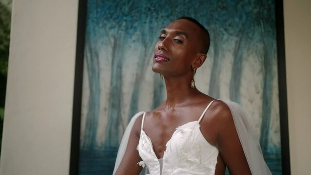 Portrait of gay black male in white wedding dress posing, smiling for fashion photo shoot. Elegant gender fluid bride sits, turns to camera at abstract blue picture in luxury retro villa. Close-up.