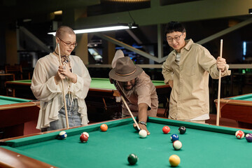 Waist up portrait of diverse group of young people playing billiards together at pool table in underground club watching friend taking shot - 782442337
