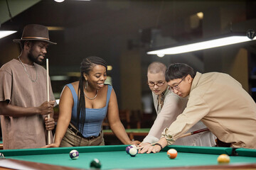 Multiethnic group of smiling young people playing pool together with Asian man taking shot at ball