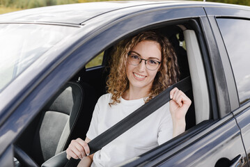 young beautiful smiling woman sitting in motionless car looking straight to camera, fastening her seatbelt, attractive caucasian woman in white t-shirt