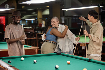 Portrait of two young women embracing at pool table enjoying game of billiards with diverse group of friends - 782444194
