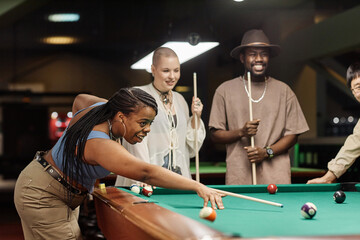 Side view portrait of smiling African American woman playing pool at table in nightclub with diverse group of friends copy space - 782444538
