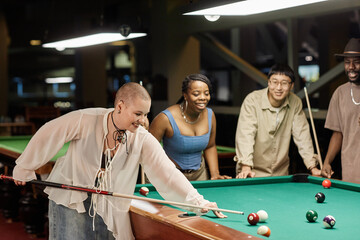 Side view portrait of smiling young bald woman playing pool at table in nightclub with diverse group of friends copy space
