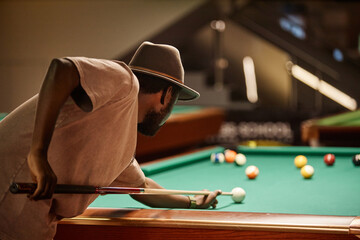 Over shoulder view of African American man playing billiards and hitting ball with cue stick in low lighting copy space