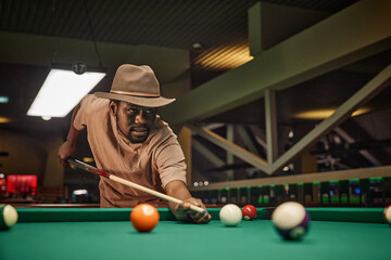 Portrait of Black young man playing billiards and wearing hat while hitting ball with cue stick in low lighting copy space