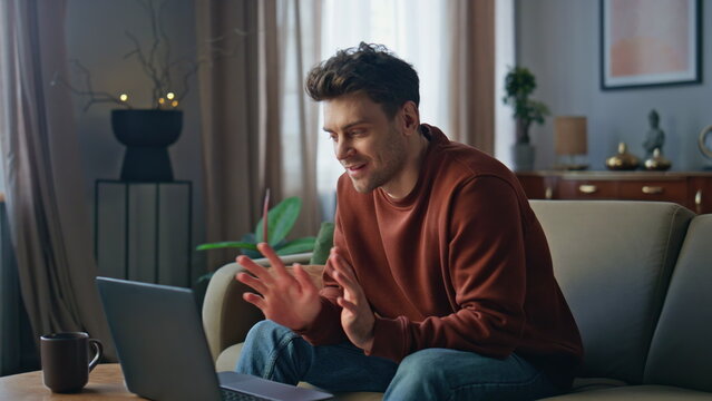 Smiling man video calling by laptop at apartment couch. Guy have online meeting