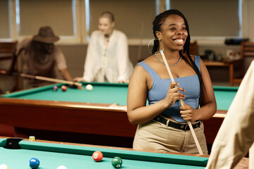 Waist up portrait of smiling African American woman chatting with friend while enjoying game of pool together in low light copy space - 782452142