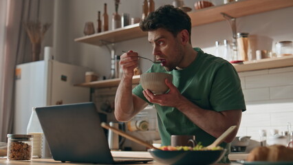 Focused man having breakfast reading laptop kitchen closeup. Guy eating granola