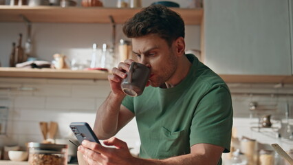 Focused man having breakfast reading cellphone at kitchen closeup. Guy drinking