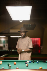 Vertical portrait of Black young man wearing leather hat and holding cue stick while standing by table in pool club - 782458131