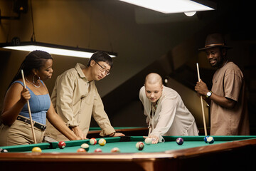 Portrait of multiethnic group of friends playing pool together by table in nightclub in muted tones