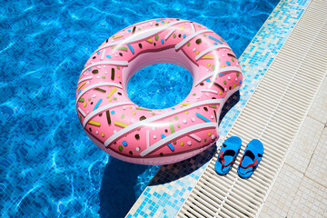 Pink inflatable ring and rubber flip-flops by blue outdoor pool water. Poolside relaxation. - obrazy, fototapety, plakaty