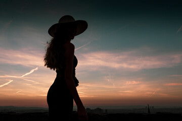 A woman wearing a hat and standing in the sunset. She is reaching out with her arms open, as if she...