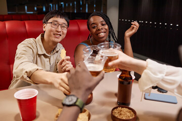 Multiethnic group of young people clinking beer bottles and cheering sitting at diner table
